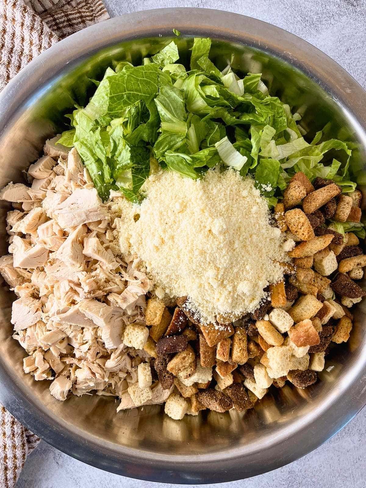 Large stainless steel bowl filled with rotini pasta topped with chopped cooked chicken, romaine lettuce, croutons, and a mound of creamy Caesar dressing with Parmesan cheese before mixing. A brown striped kitchen towel sits beside the bowl on a gray countertop.