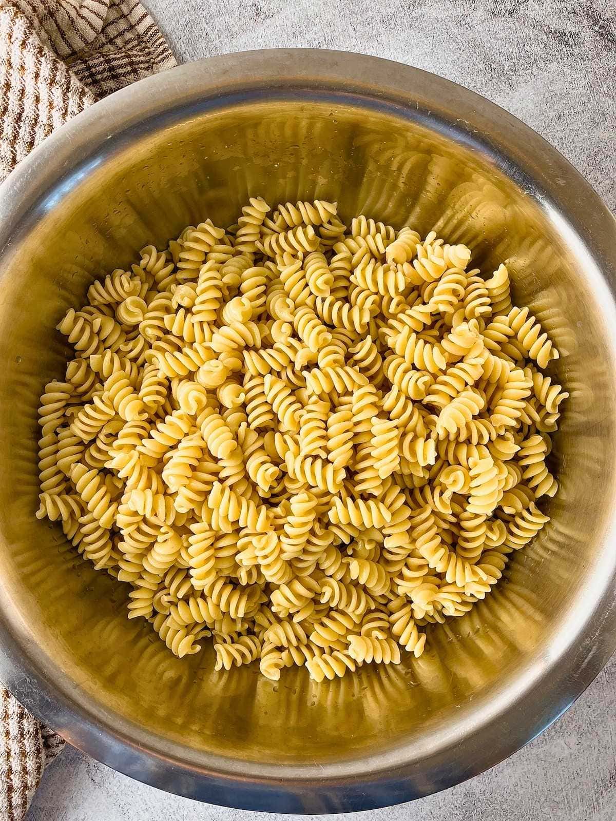Cooked rotini pasta in a large stainless steel mixing bowl on a gray countertop. A striped kitchen towel sits beside the bowl.