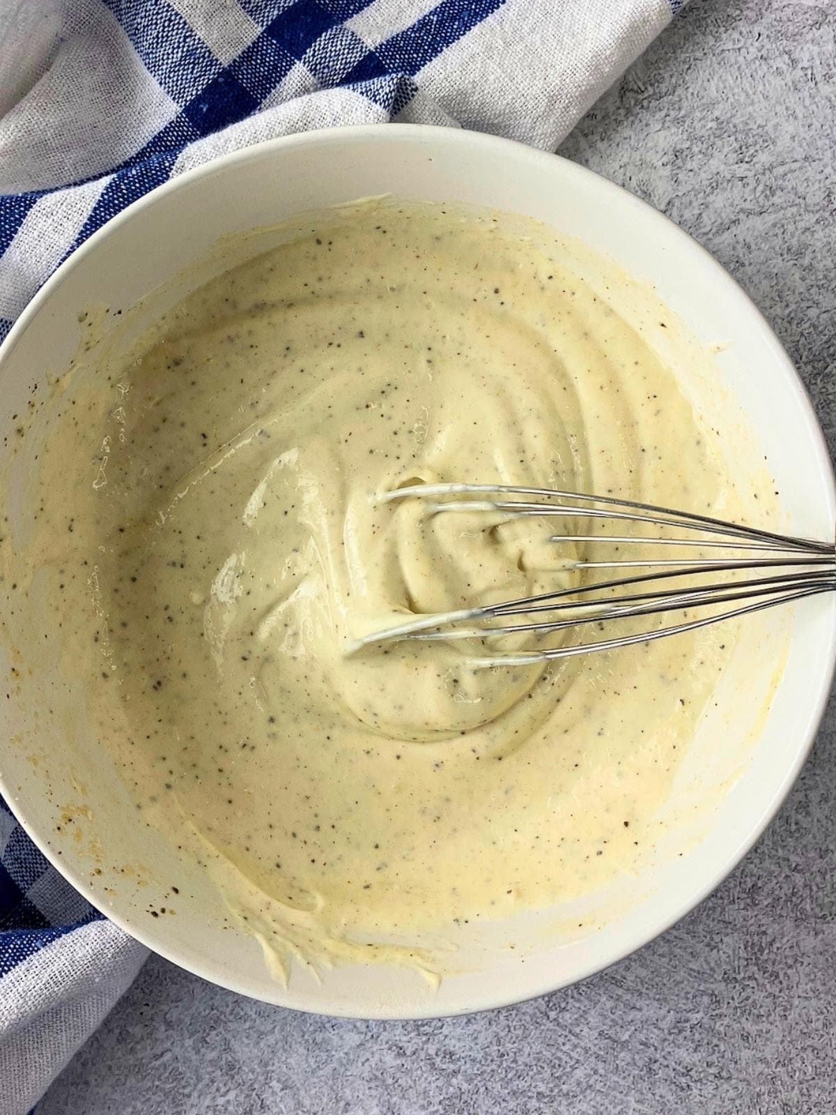 Caesar dressing whisked together in a white bowl until smooth and creamy with black pepper speckled throughout. A metal whisk rests in the bowl on a gray countertop beside a blue and white checkered towel.