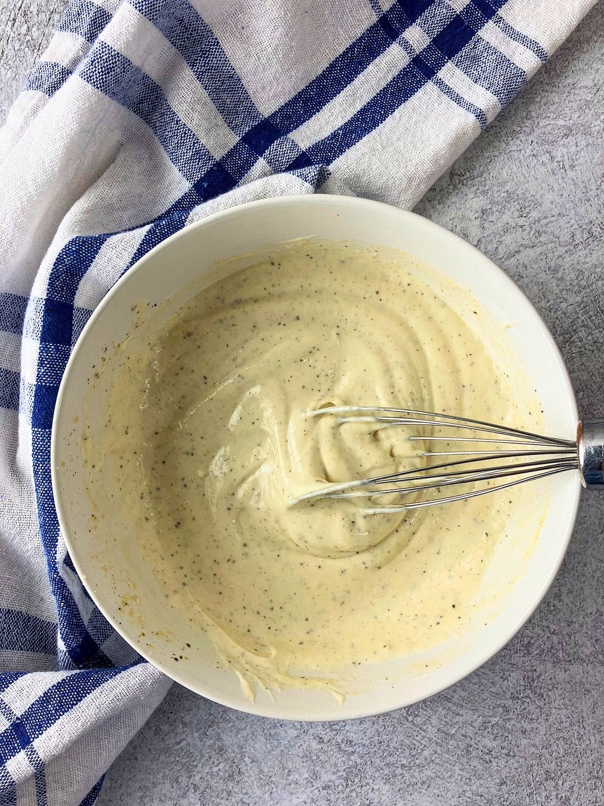 Seasoned Caesar dressing is whisked together in a white bowl until smooth and speckled with black pepper. A metal whisk rests in the creamy mixture beside a blue and white checkered towel.