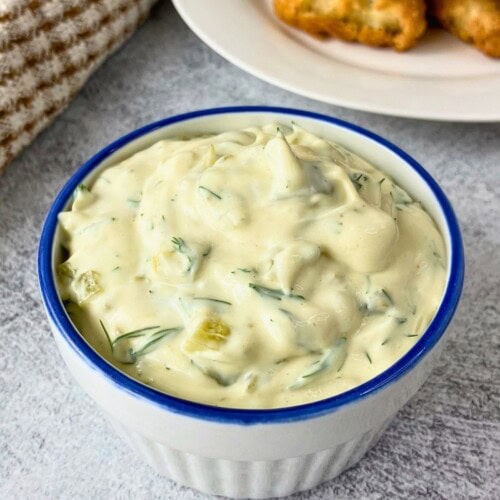 White bowl filled with easy tartar sauce recipe made with creamy sauce, dill, and chopped pickle pieces, with fried fish on a plate in the background.