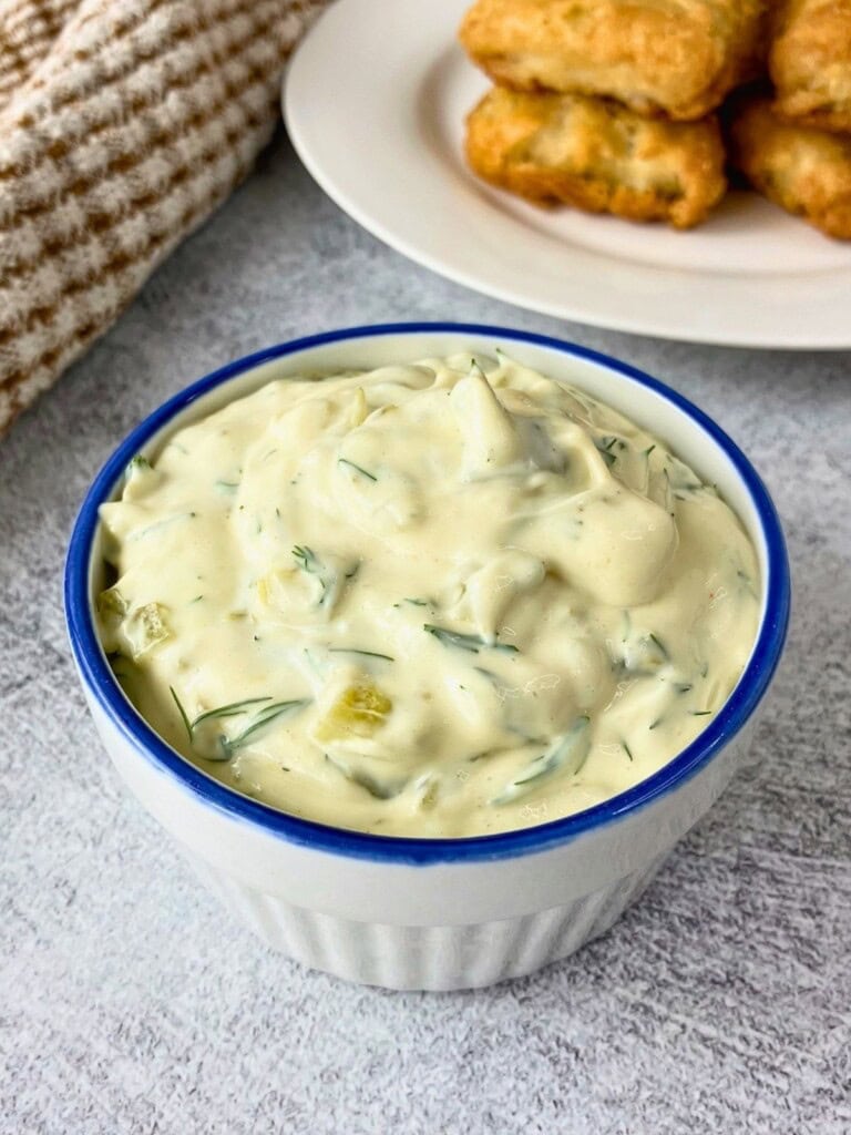 White bowl filled with easy tartar sauce recipe made with creamy sauce, dill, and chopped pickle pieces, with fried fish on a plate in the background.