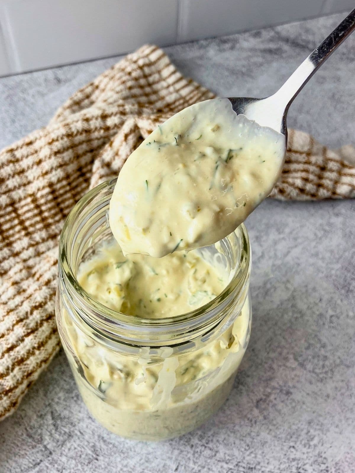 Spoonful of creamy tartar sauce with dill and chopped pickle pieces lifted from a glass jar for an easy tartar sauce recipe.