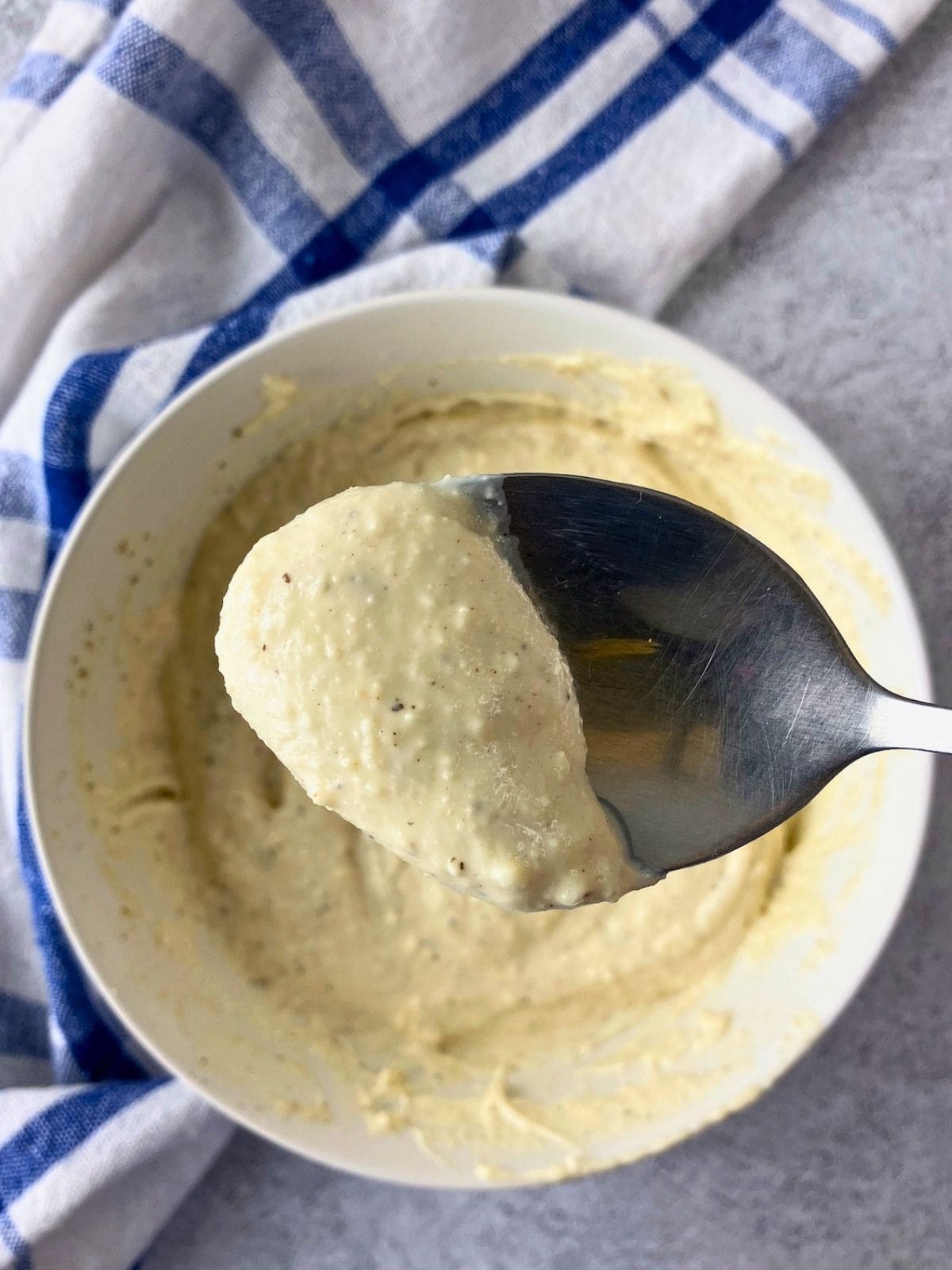 Spoonful of thick homemade Caesar dressing held over a white bowl of creamy dressing. The dressing is speckled with black pepper and Parmesan cheese.