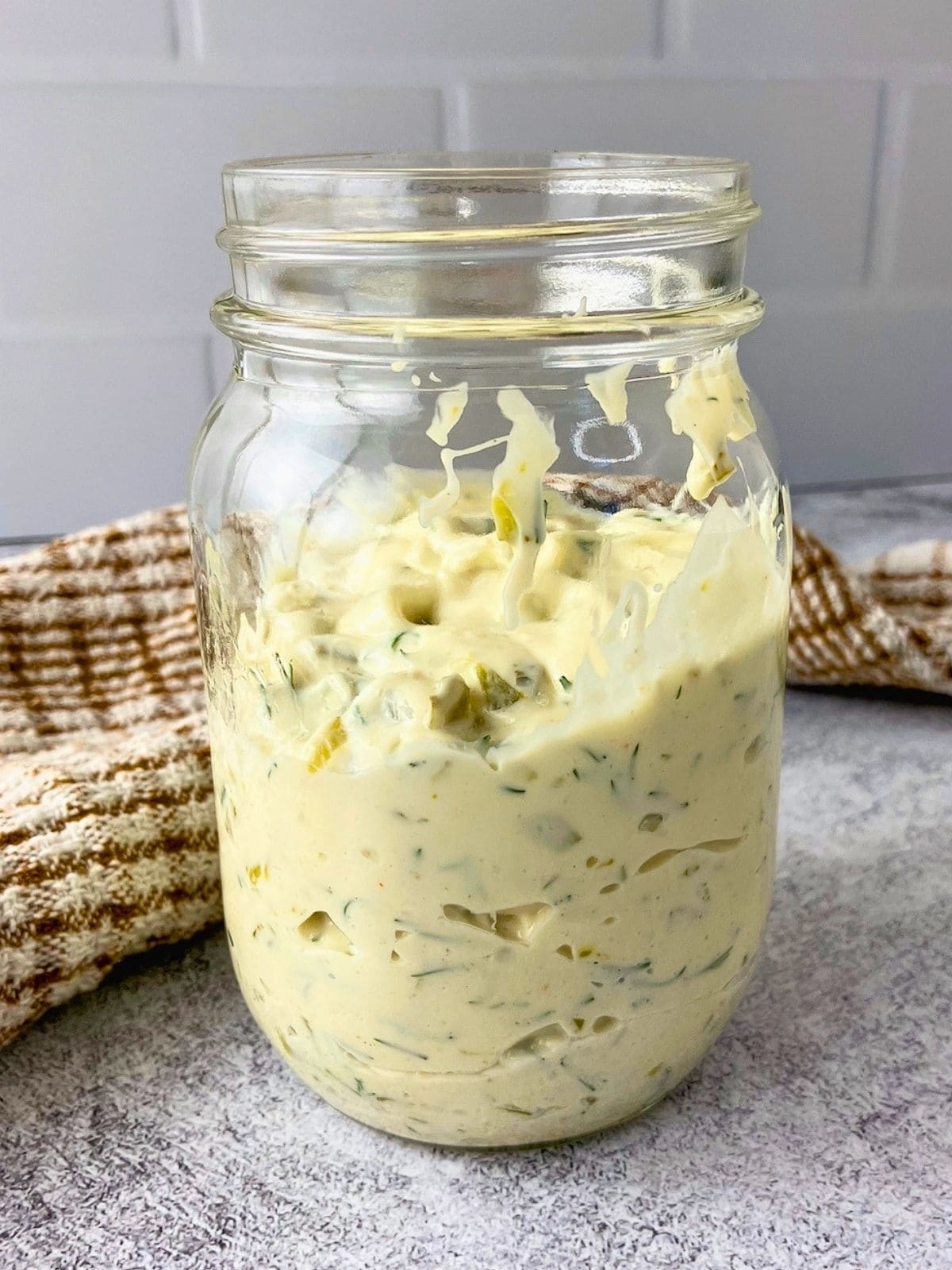 Glass jar filled with creamy tartar sauce with dill and chopped pickle pieces on a gray counter for an easy tartar sauce recipe.