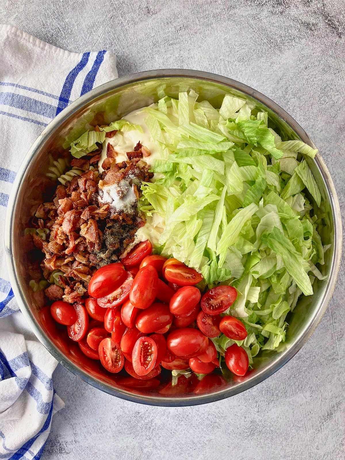 Sliced grape tomatoes, crispy bacon, shredded lettuce, and seasoning are added to rotini pasta and Miracle Whip in a stainless steel bowl for BLT pasta salad.