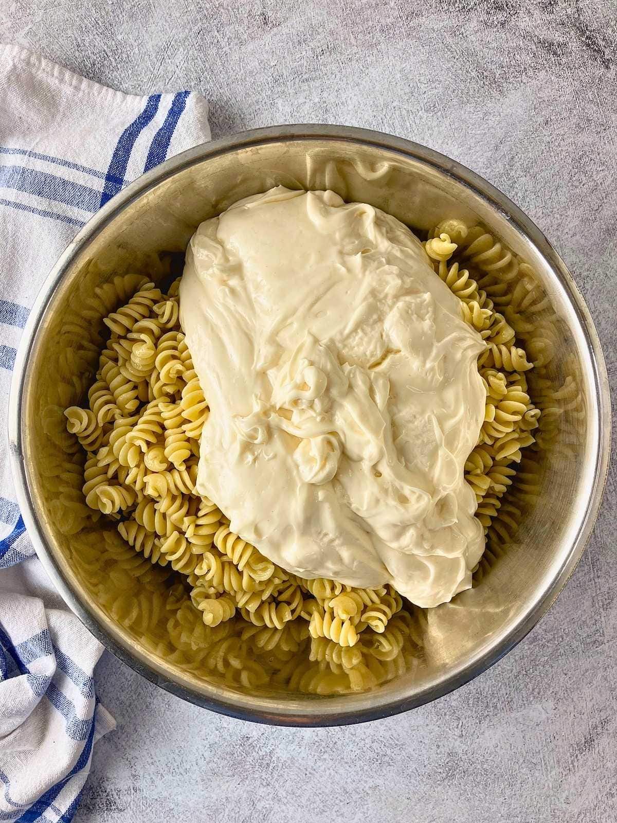 A large scoop of Miracle Whip sits on top of cooked rotini pasta in a stainless steel mixing bowl as the creamy base for the pasta salad.