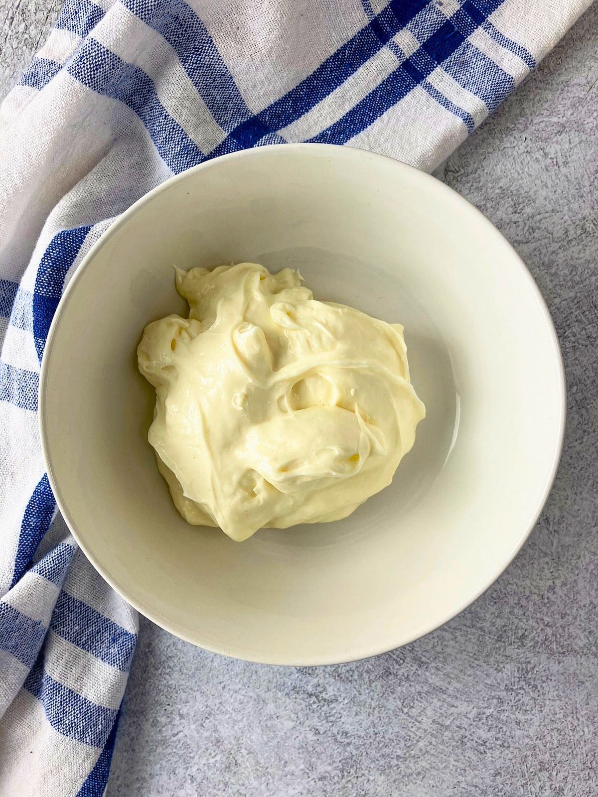 A scoop of Miracle Whip in a white bowl on a gray countertop. A blue and white checkered towel sits beside the bowl.