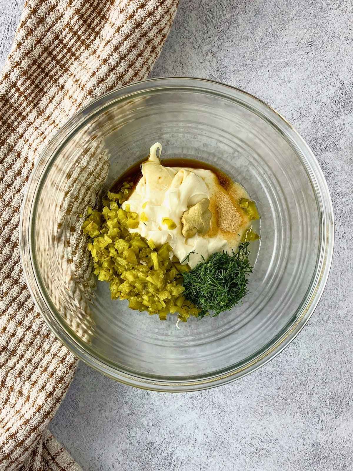 Glass bowl with mayonnaise, chopped pickles, fresh dill, mustard, and seasonings ready to be mixed.