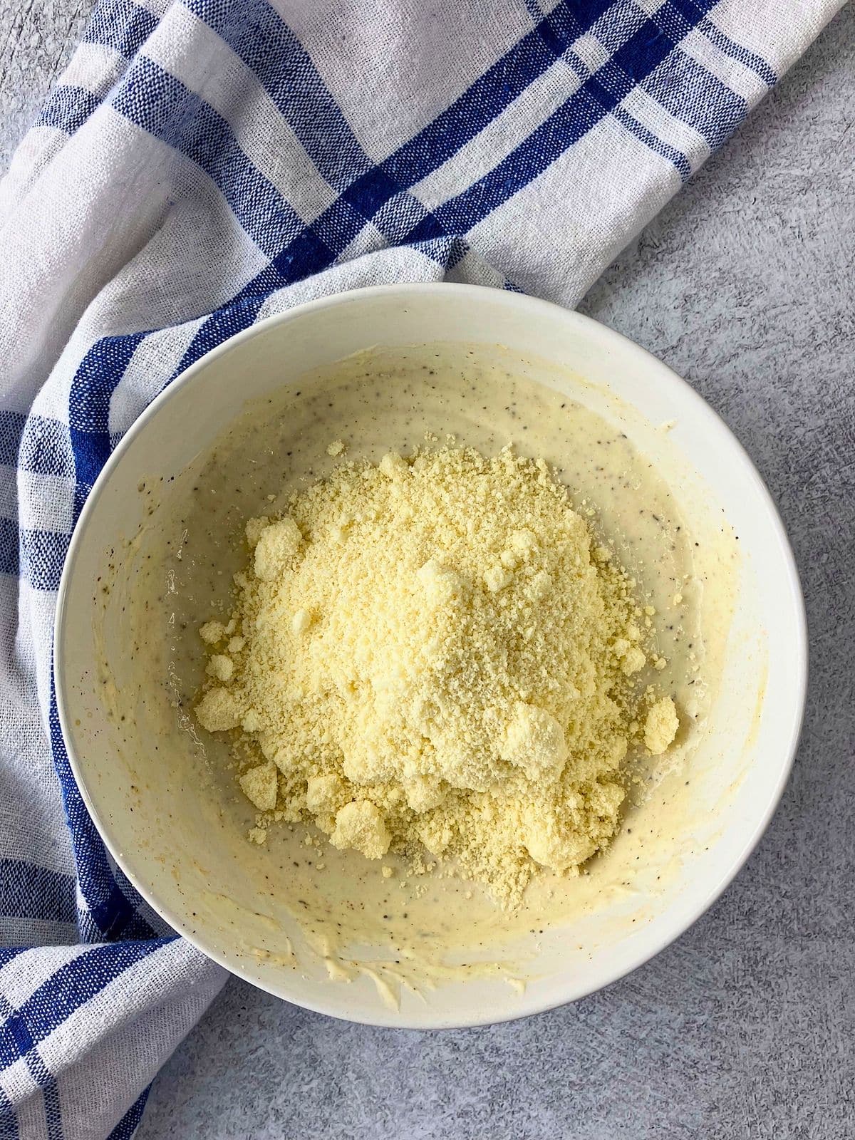 Grated Parmesan cheese added to a bowl of creamy seasoned dressing. The white bowl sits on a gray countertop beside a blue and white checkered towel.