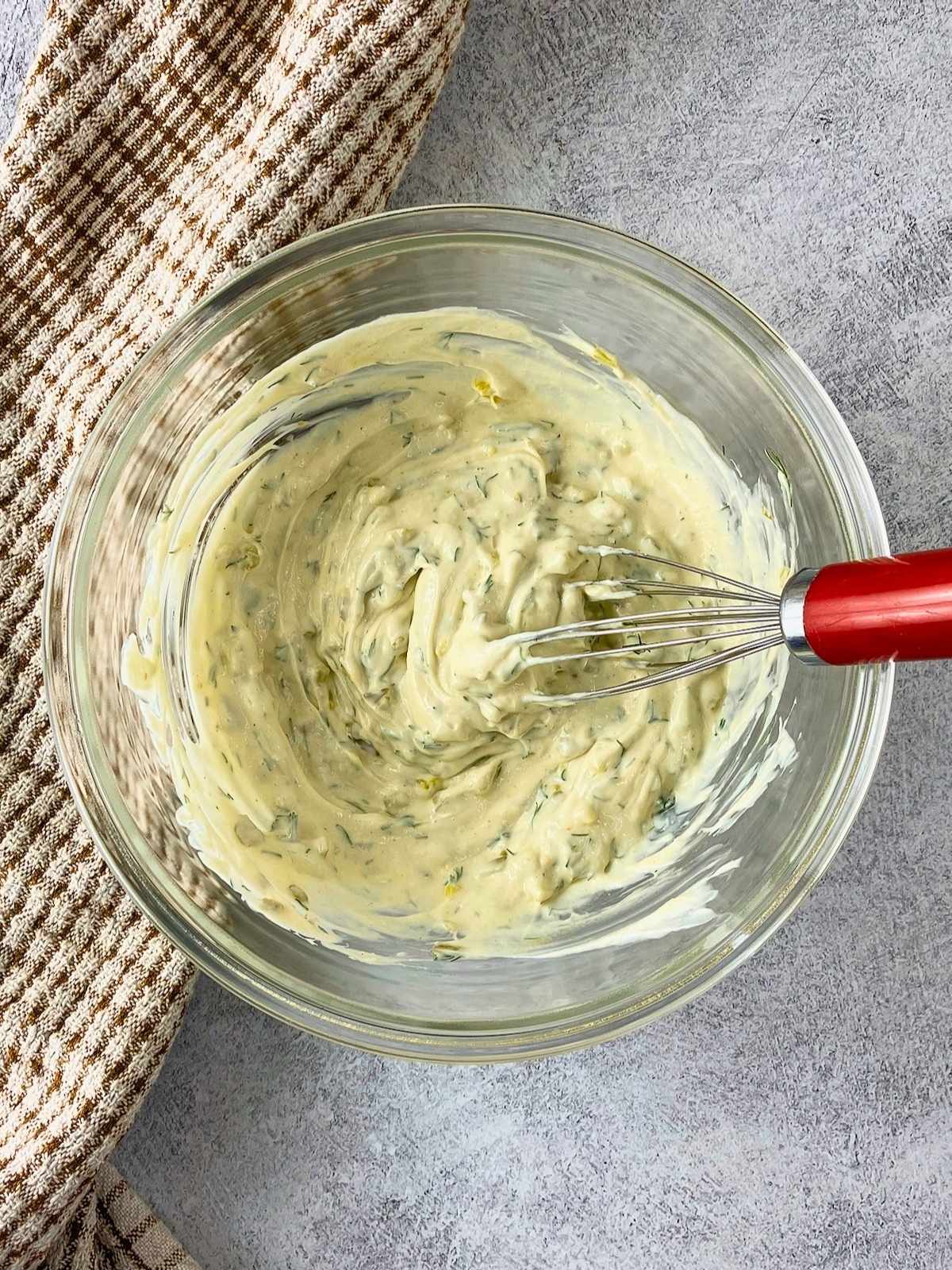 Glass bowl of creamy tartar sauce with dill and chopped pickles being mixed with a red handled whisk.