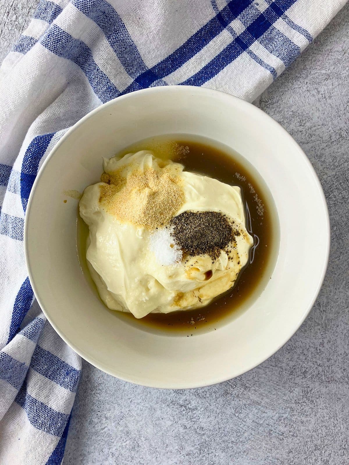 Miracle Whip in a white bowl topped with seasonings and Worcestershire sauce for mixing into a sauce or dressing. A blue and white checkered towel sits beside the bowl on a gray countertop.