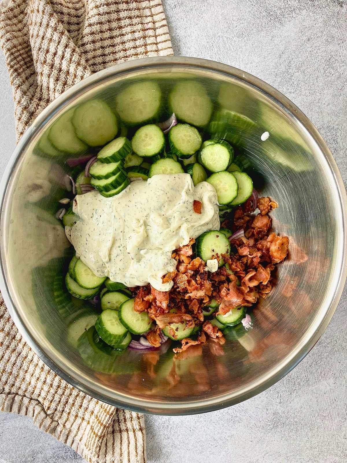 Sliced cucumbers, red onion, bacon pieces, and creamy dill dressing in a mixing bowl.