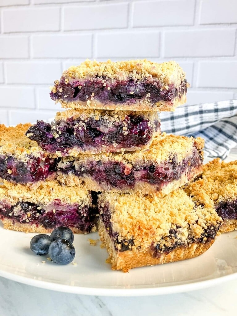 Blueberry crumb bars stacked on a white plate with fresh blueberries in front.