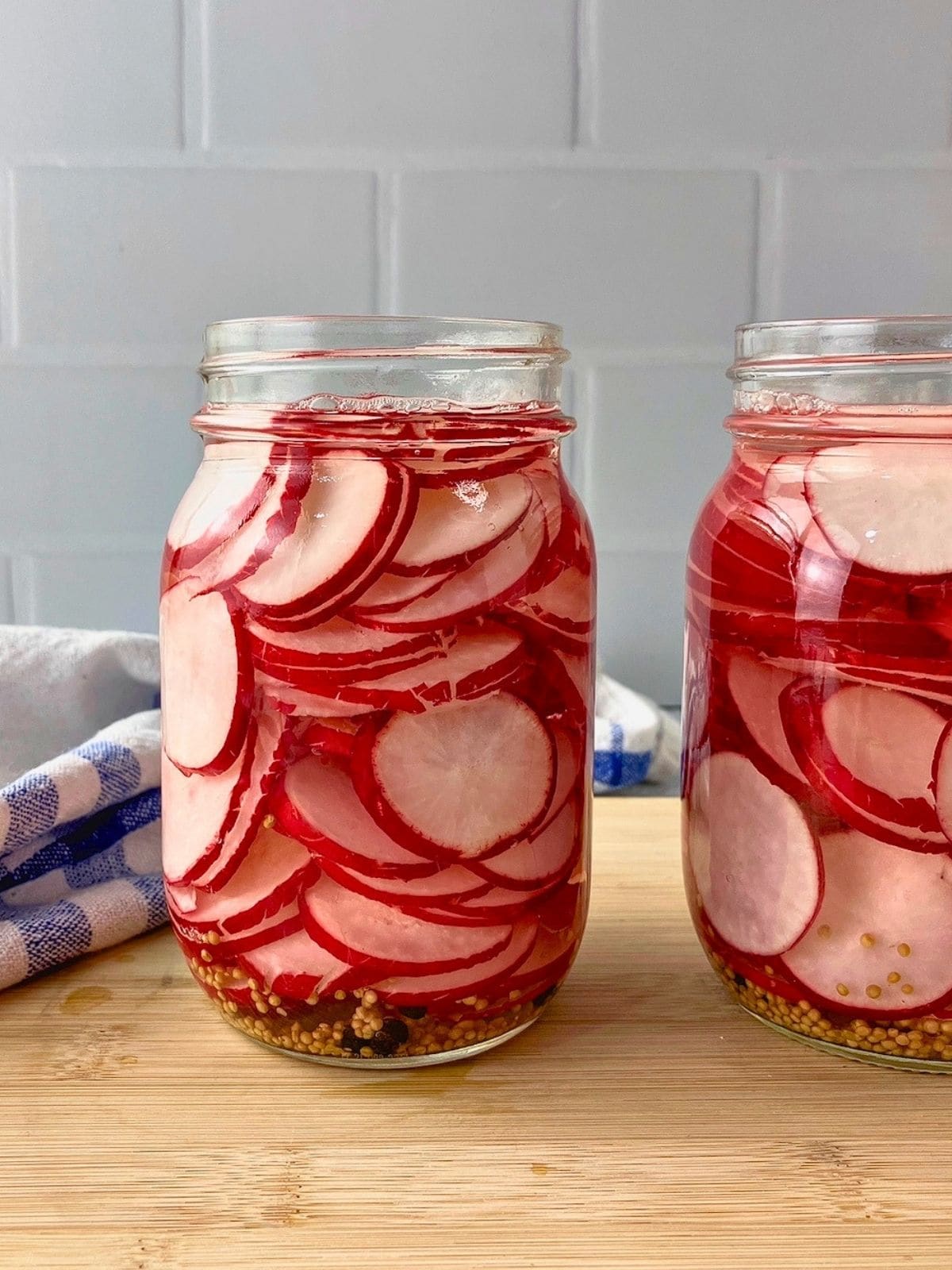 Two glass jars filled with thin radish slices in red pickling brine, with mustard seeds and peppercorns at the bottom.