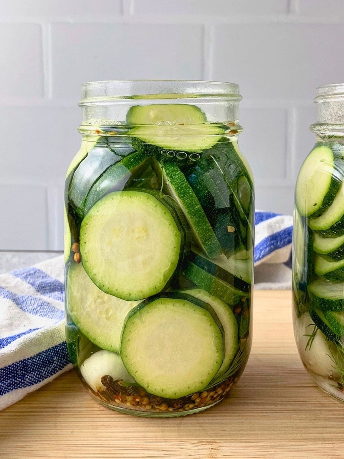 Jars of zucchini dill pickles with sliced zucchini, garlic, dill, peppercorns, pickling spice, and red pepper flakes, with the brine poured over top.