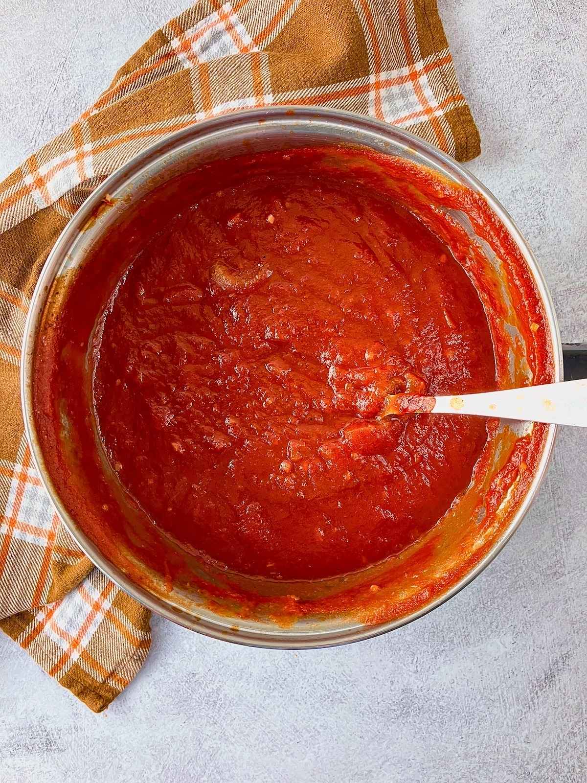 Pot of thick red homemade ketchup being stirred with a spoon on a gray counter beside an orange plaid towel.