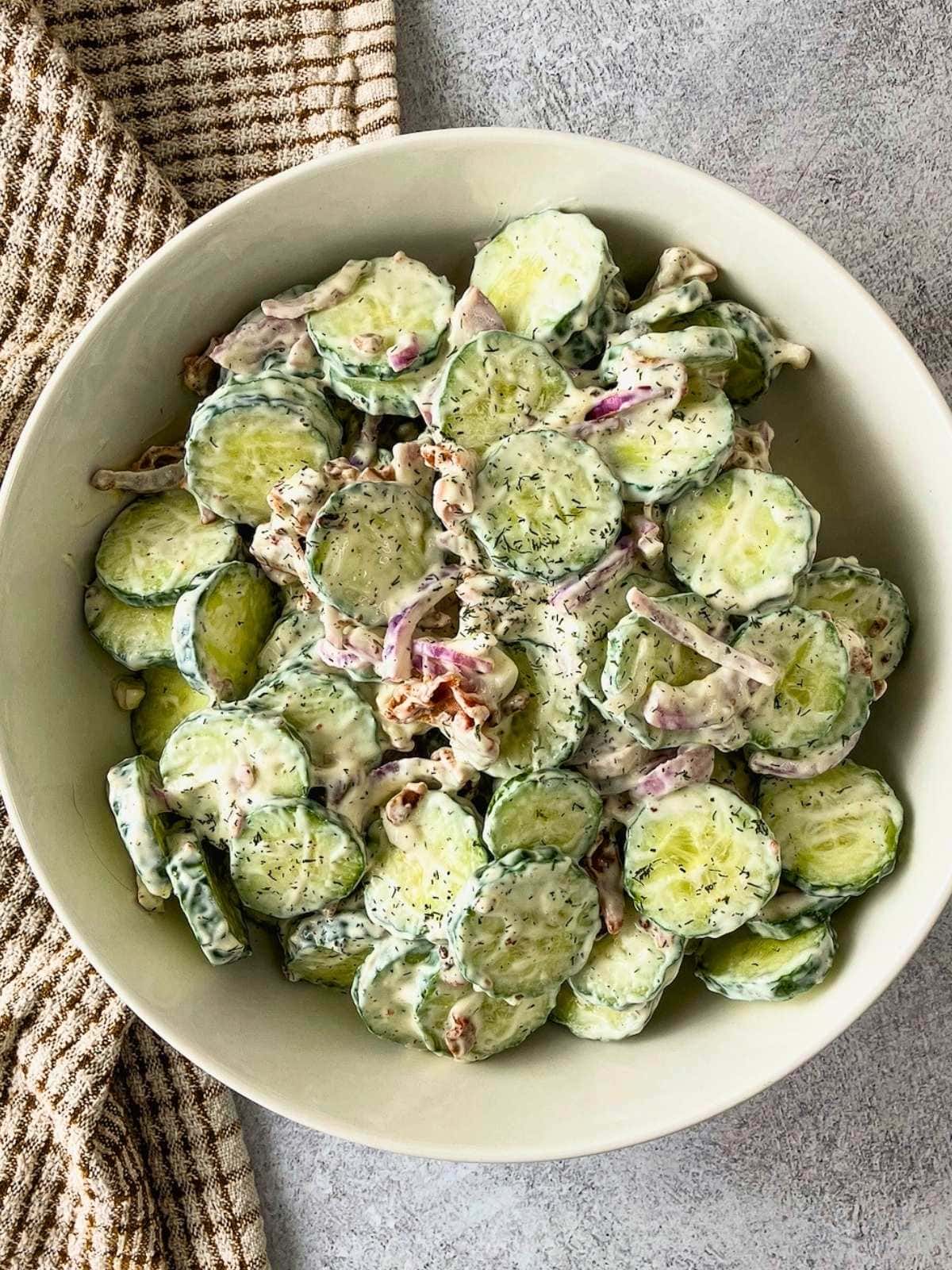 Creamy bacon cucumber salad with sliced cucumbers, red onion, bacon pieces, and dill served in a white bowl.