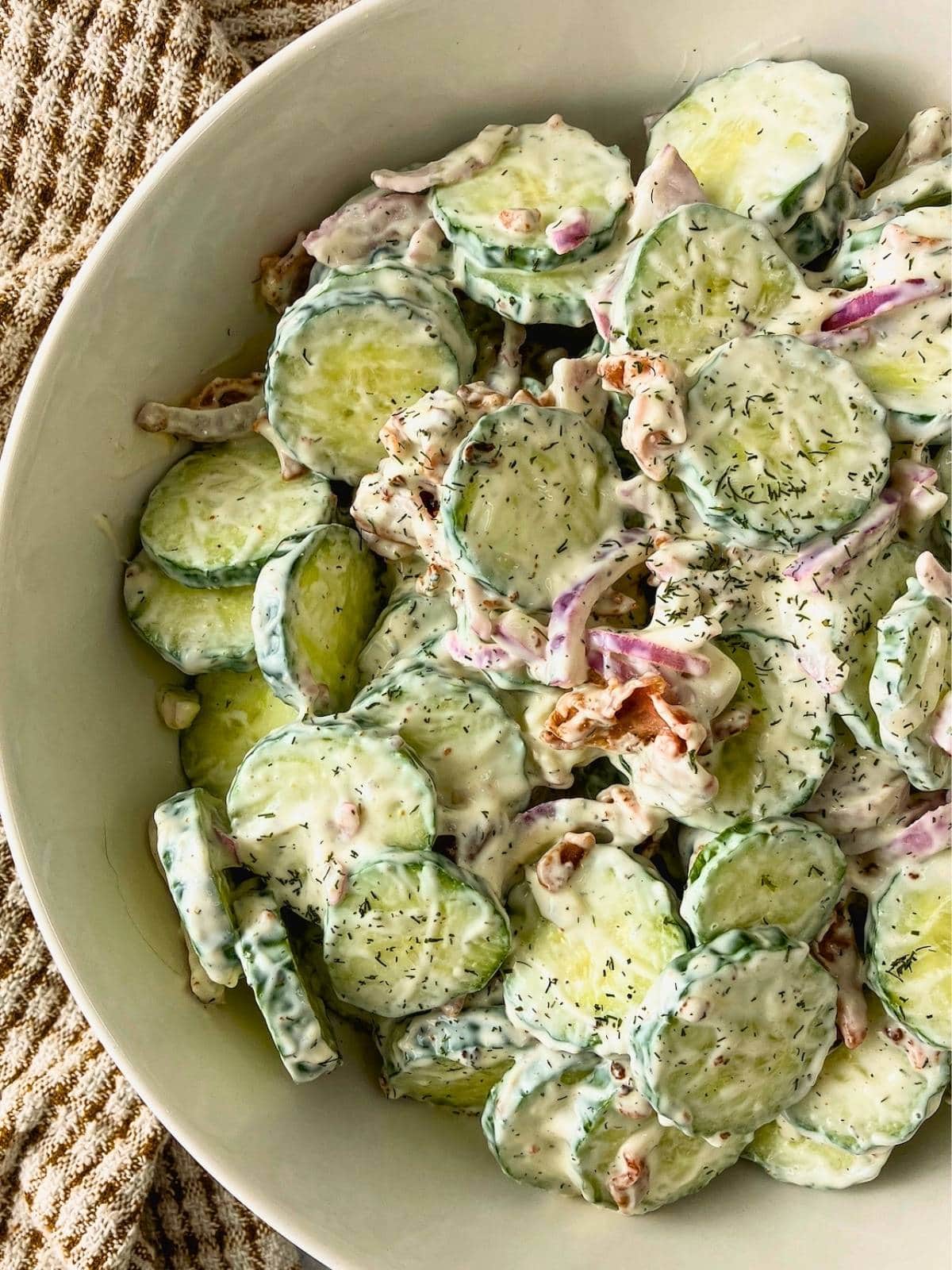 Creamy bacon cucumber salad with sliced cucumbers, red onion, bacon pieces, and dill in a white bowl.