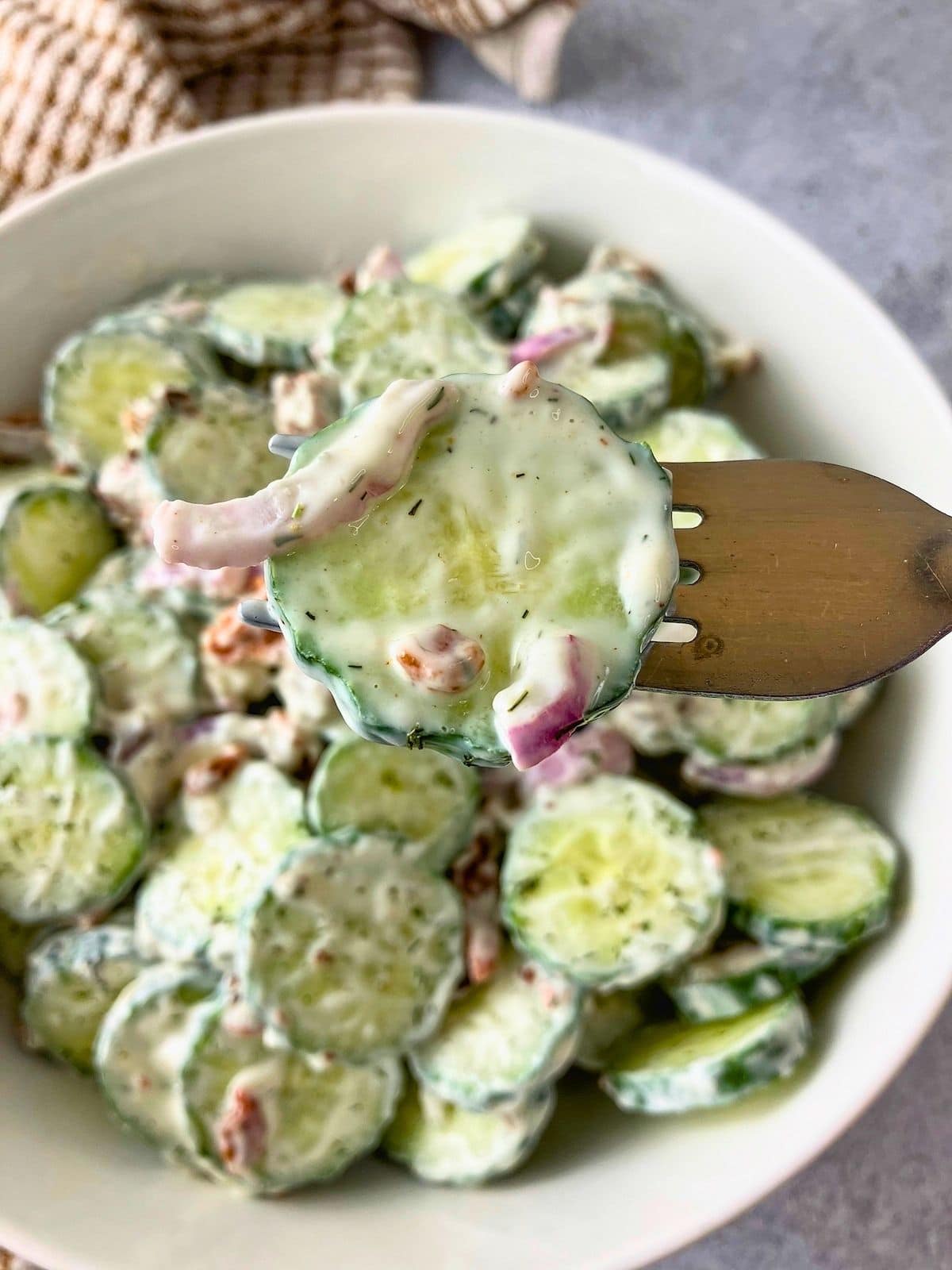 Creamy bacon cucumber salad served in a white bowl, with sliced cucumbers, red onion, and bacon in a creamy dressing and a fork lifting some up.