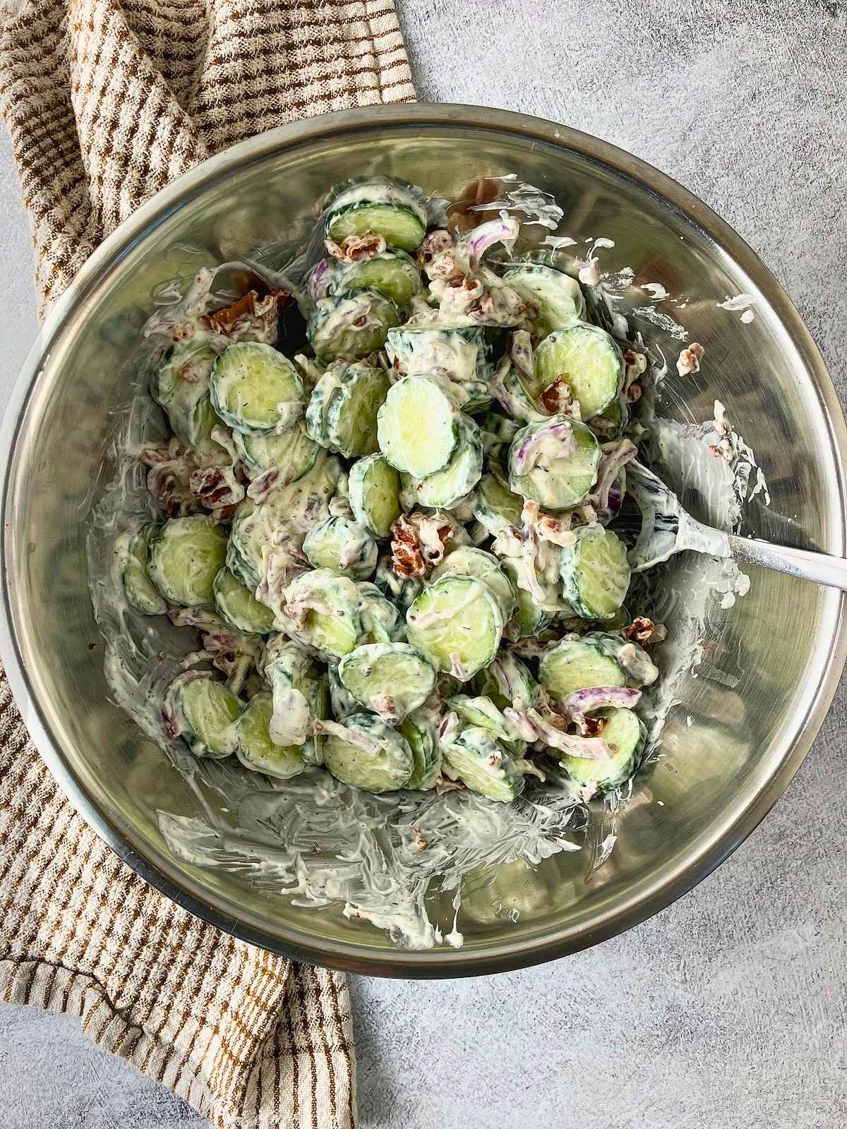 Creamy cucumber salad with sliced cucumbers, red onion, and bacon pieces tossed in a mixing bowl.