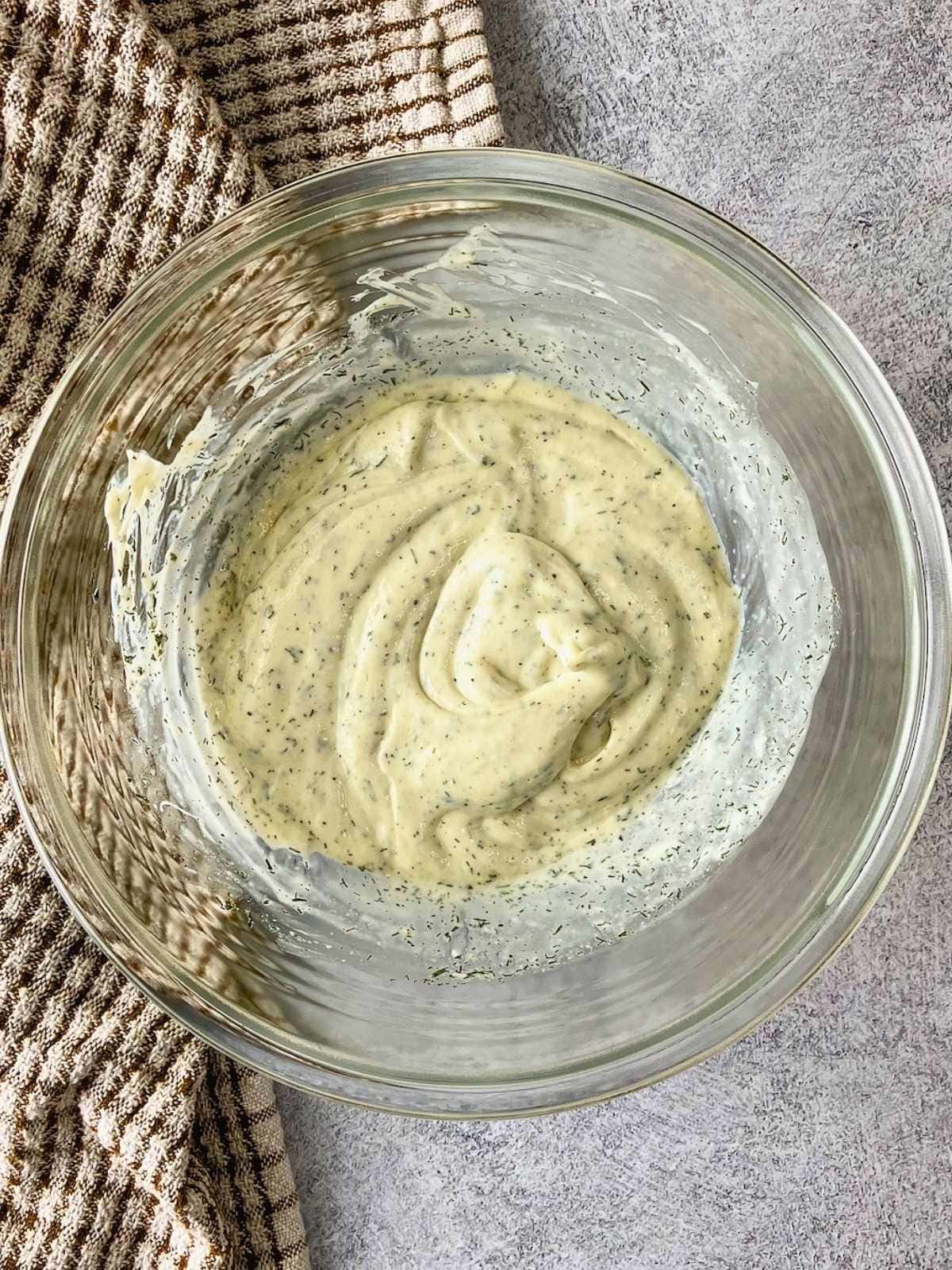 Creamy cucumber salad dressing mixed with dill and seasonings in a glass bowl.