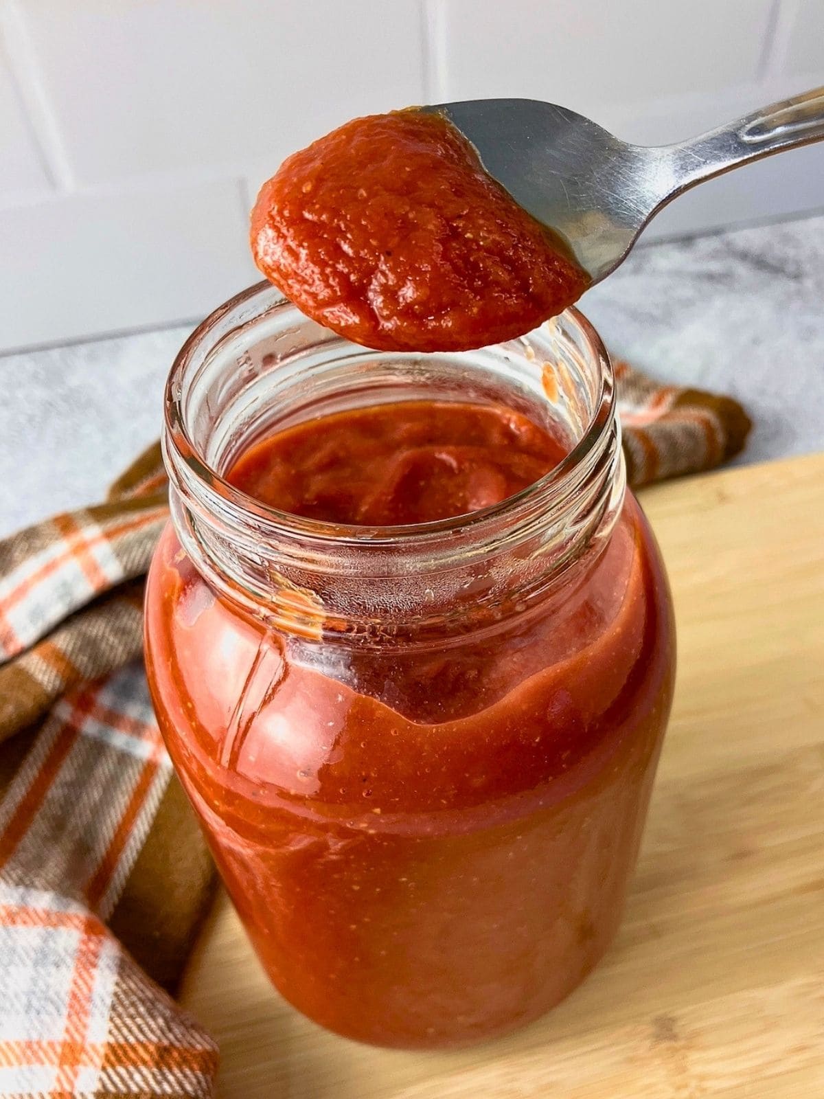 A spoon lifts thick easy homemade ketchup from a glass jar on a wooden board.