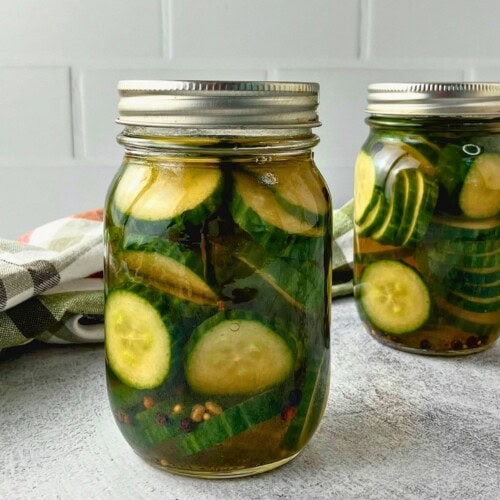 Two jars of sliced cucumber refrigerator pickles in brine with peppercorns, pickling spice, and celery seed.