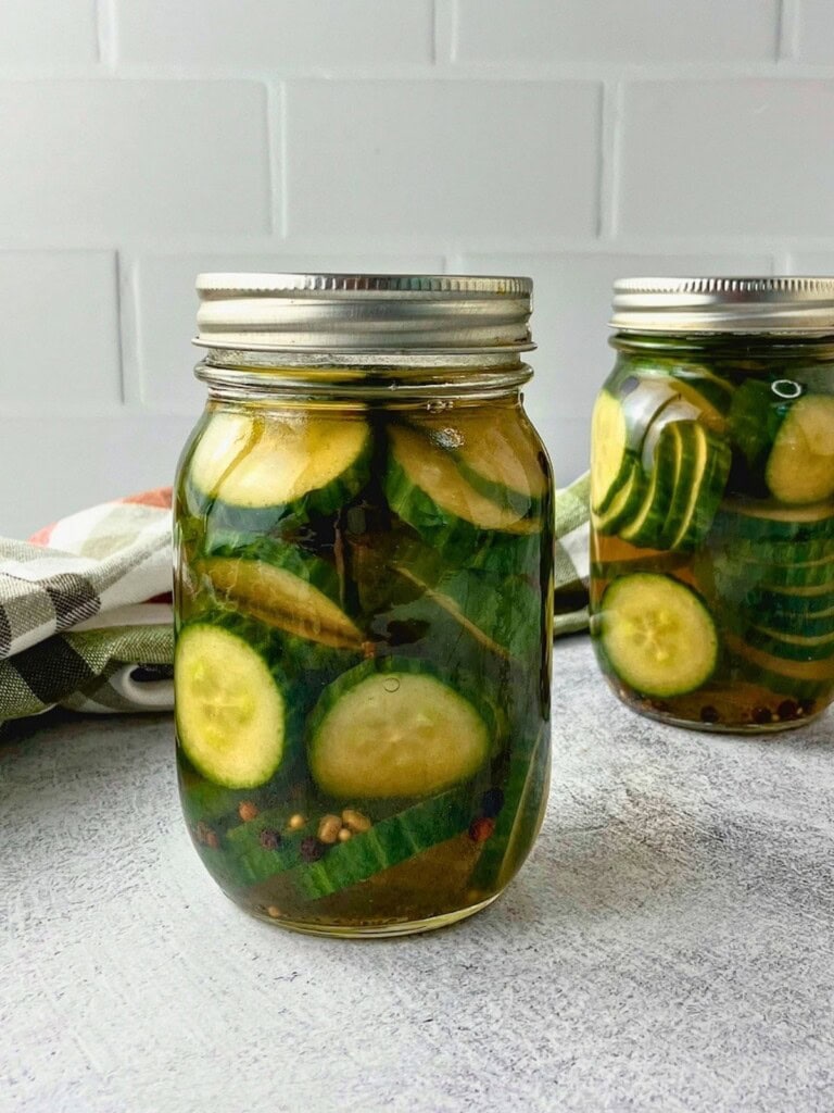 Two jars of sliced cucumber refrigerator pickles in brine with peppercorns, pickling spice, and celery seed.