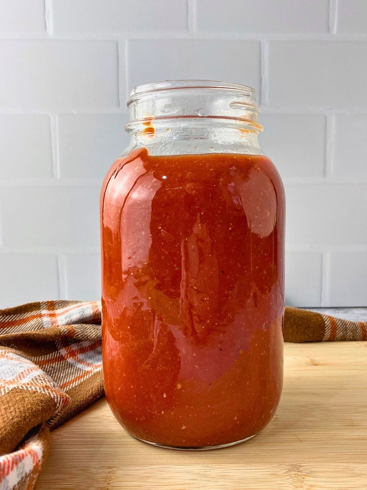 A large glass jar holds thick red homemade ketchup on a wooden board beside an orange plaid towel.