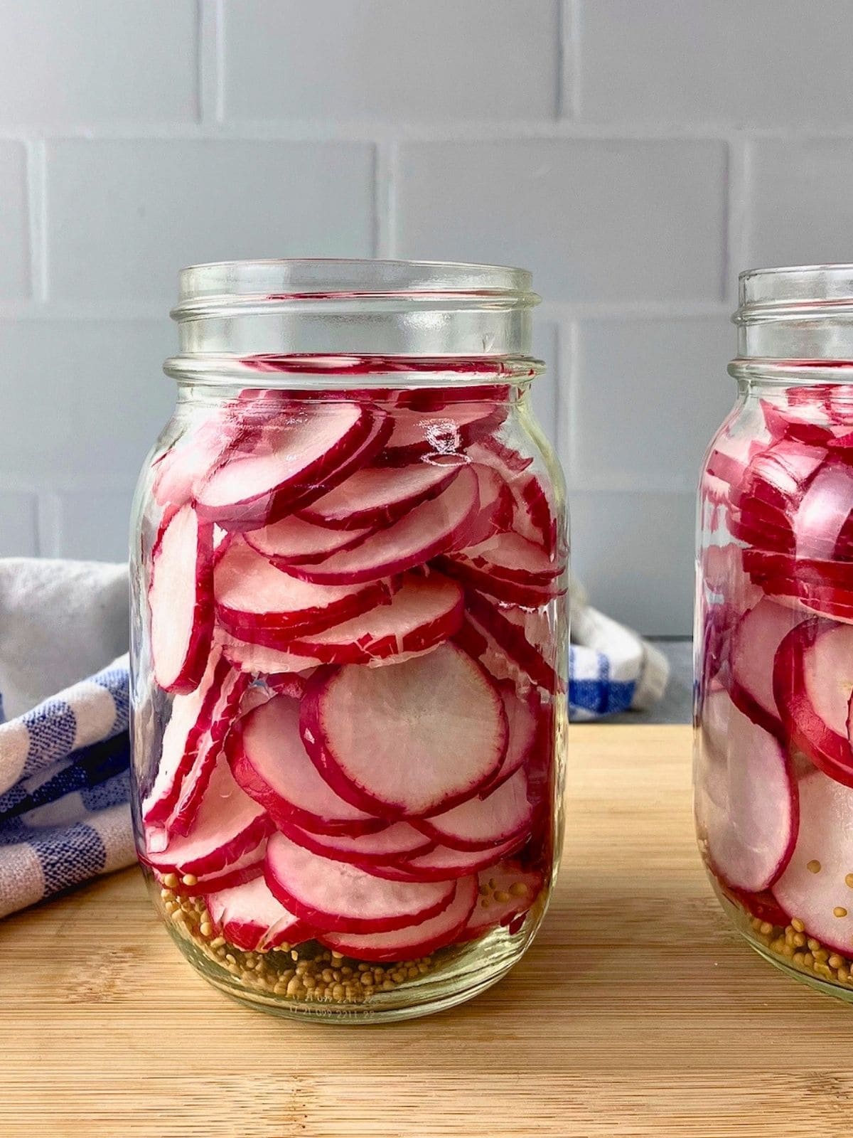 Glass jars filled with thin red radish slices and mustard seeds before the pickling brine is added.
