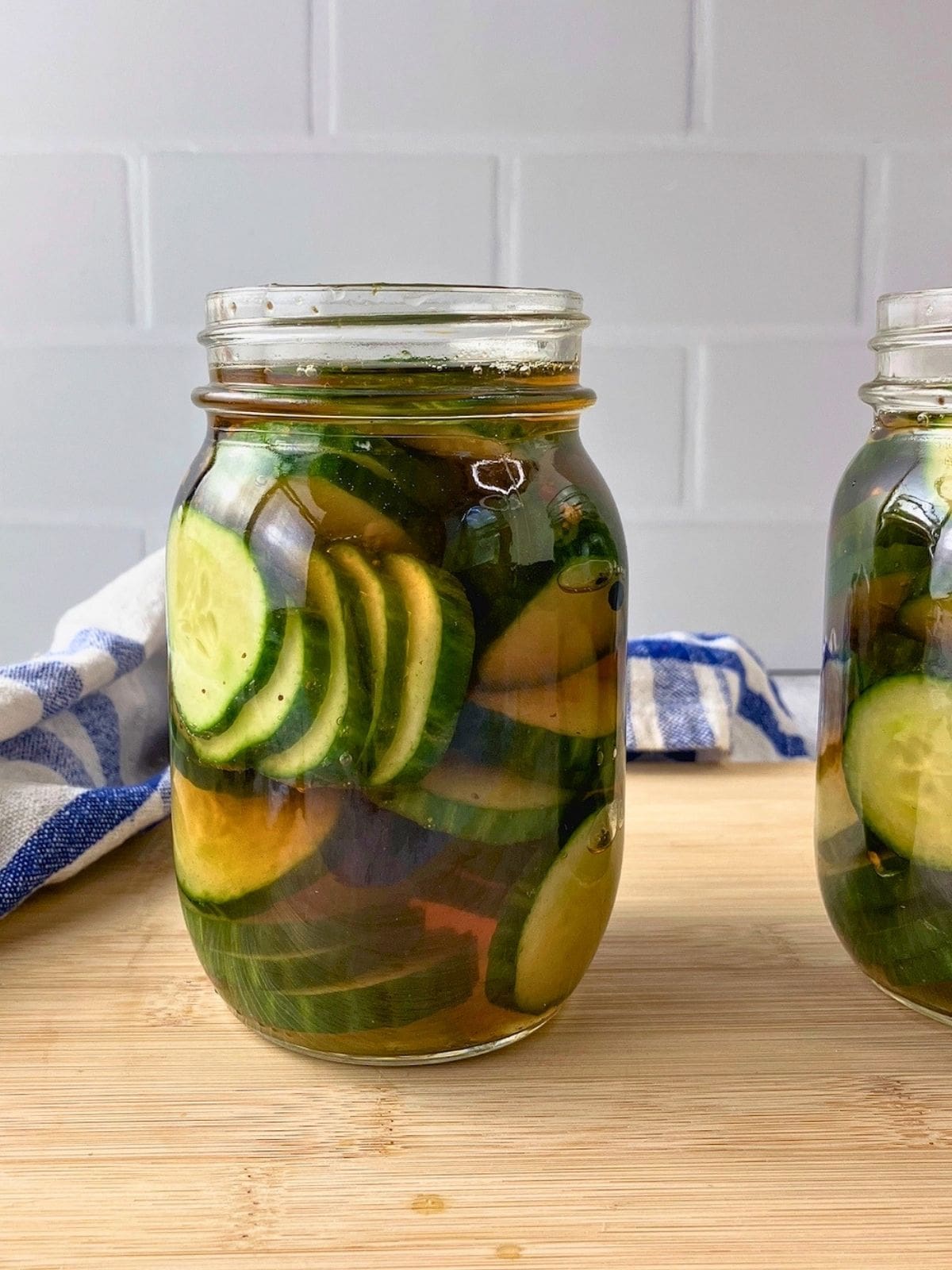 Glass jar filled with sliced refrigerator pickles in brine with peppercorns, pickling spice, and celery seed cooling on the counter.