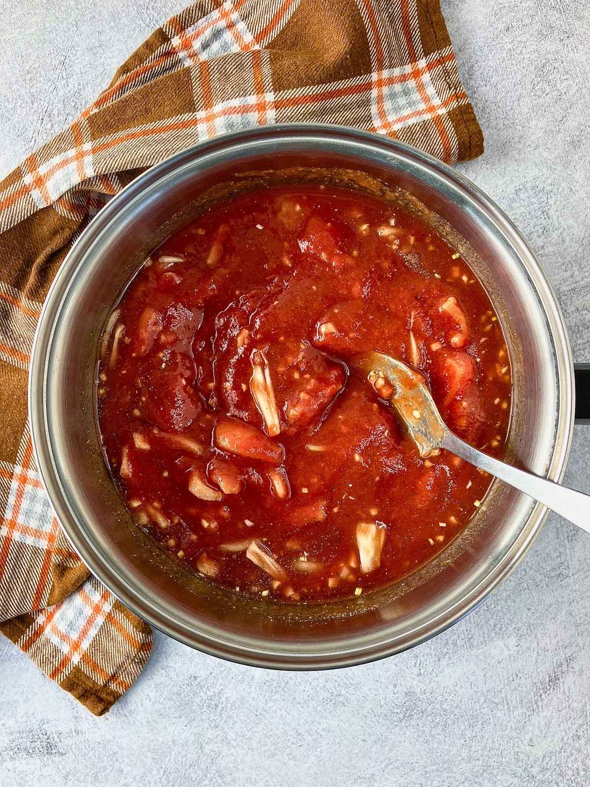Pot of tomato mixture with chopped onion and garlic being stirred with a spoon.