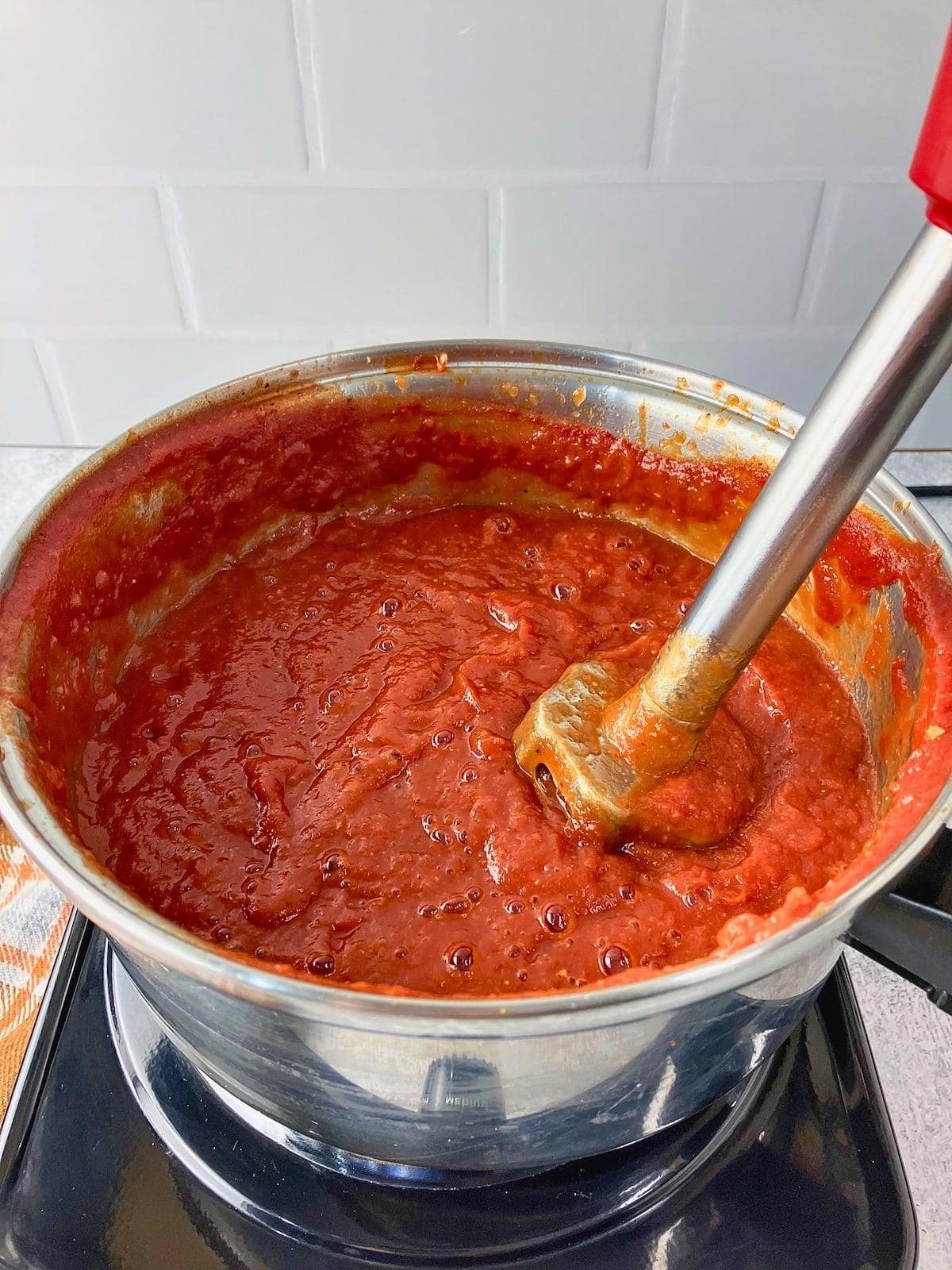 Homemade ketchup in a pot while an immersion blender smooths the thick red sauce.