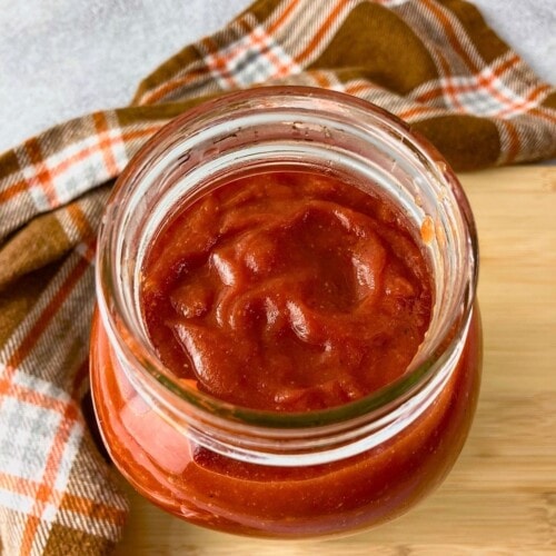Open glass jar of thick homemade ketchup on a wooden board beside an orange plaid towel.