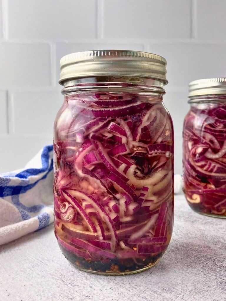 Glass jar filled with sliced pickled red onions in pale brine with peppercorns and seasonings at the bottom.