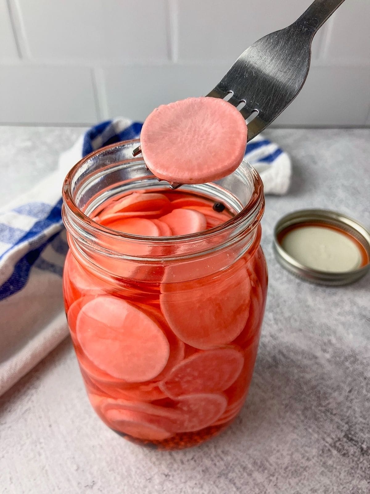 Fork lifting a pink pickled radish slice from a glass jar filled with red brine and thin radish rounds.