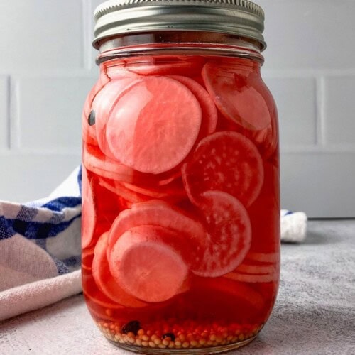 Glass jar of pickled radish slices in red brine with mustard seeds and peppercorns settled at the bottom.