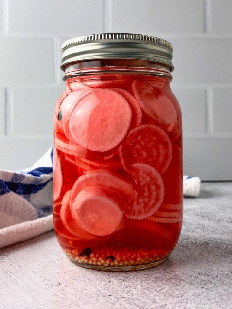 Glass jar of pickled radish slices in red brine with mustard seeds and peppercorns settled at the bottom.