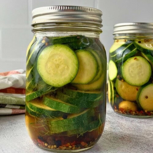 Two jars of refrigerator zucchini dill pickles with sliced zucchini, dill, garlic, peppercorns, pickling spice, and red pepper flakes in brine.