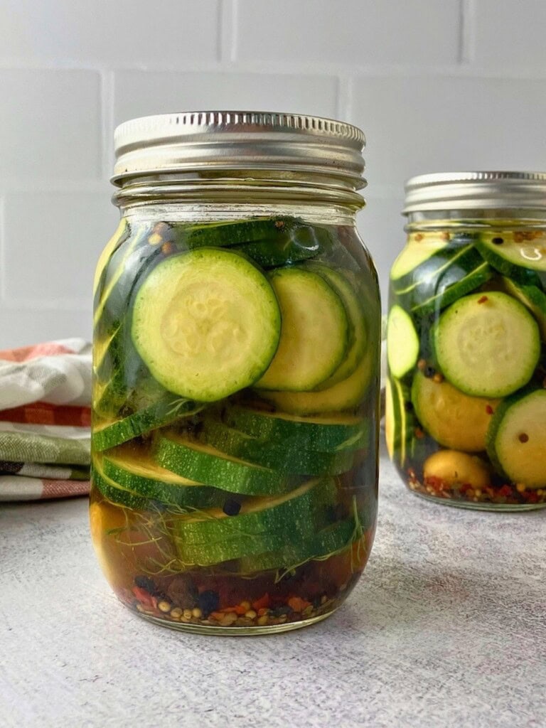 Two jars of refrigerator zucchini dill pickles with sliced zucchini, dill, garlic, peppercorns, pickling spice, and red pepper flakes in brine.