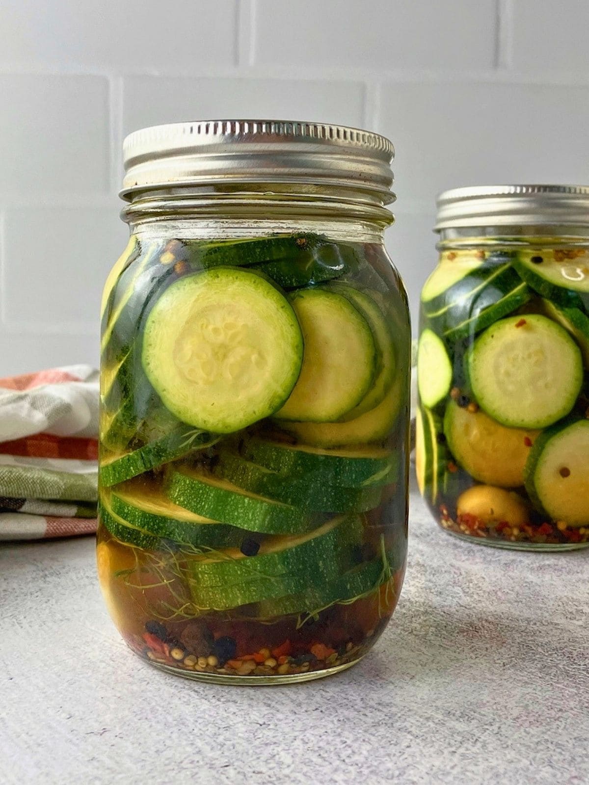 Two jars of refrigerator zucchini dill pickles with sliced zucchini, dill, garlic, peppercorns, pickling spice, and red pepper flakes in brine.