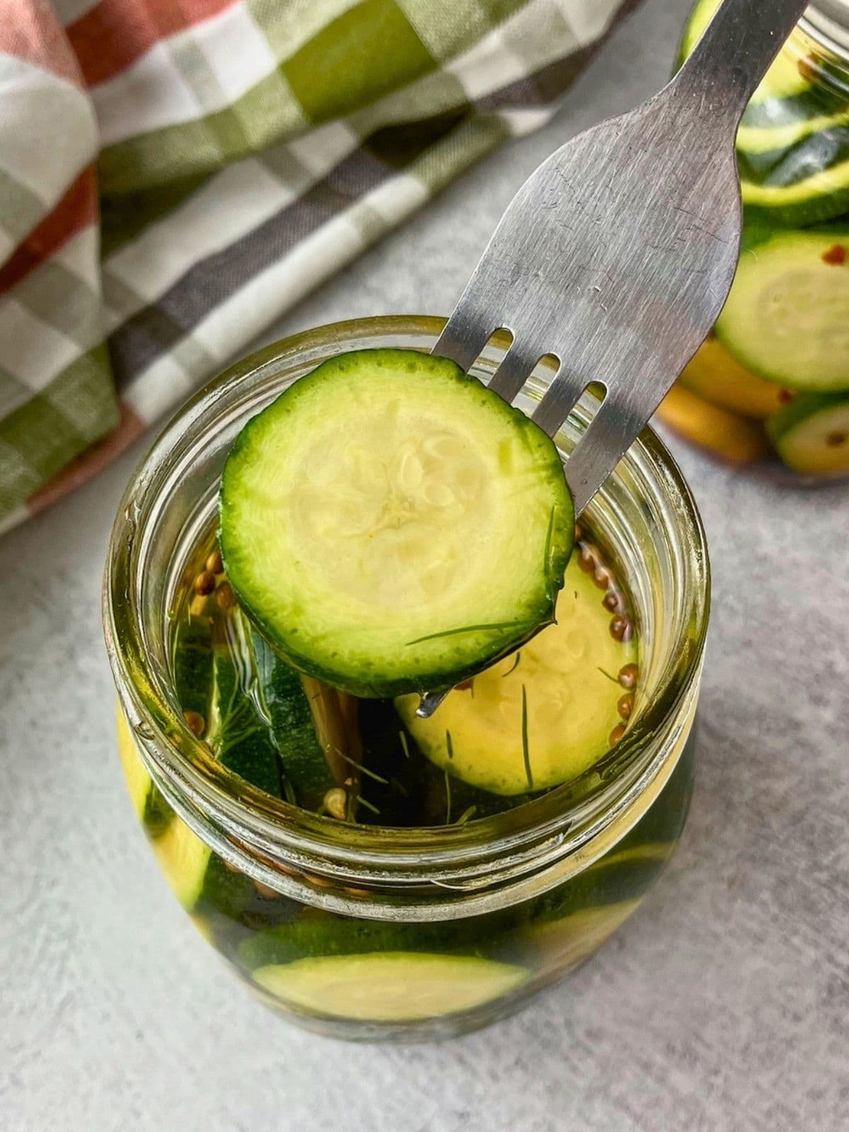 A fork lifting a slice of zucchini dill pickle from a jar.