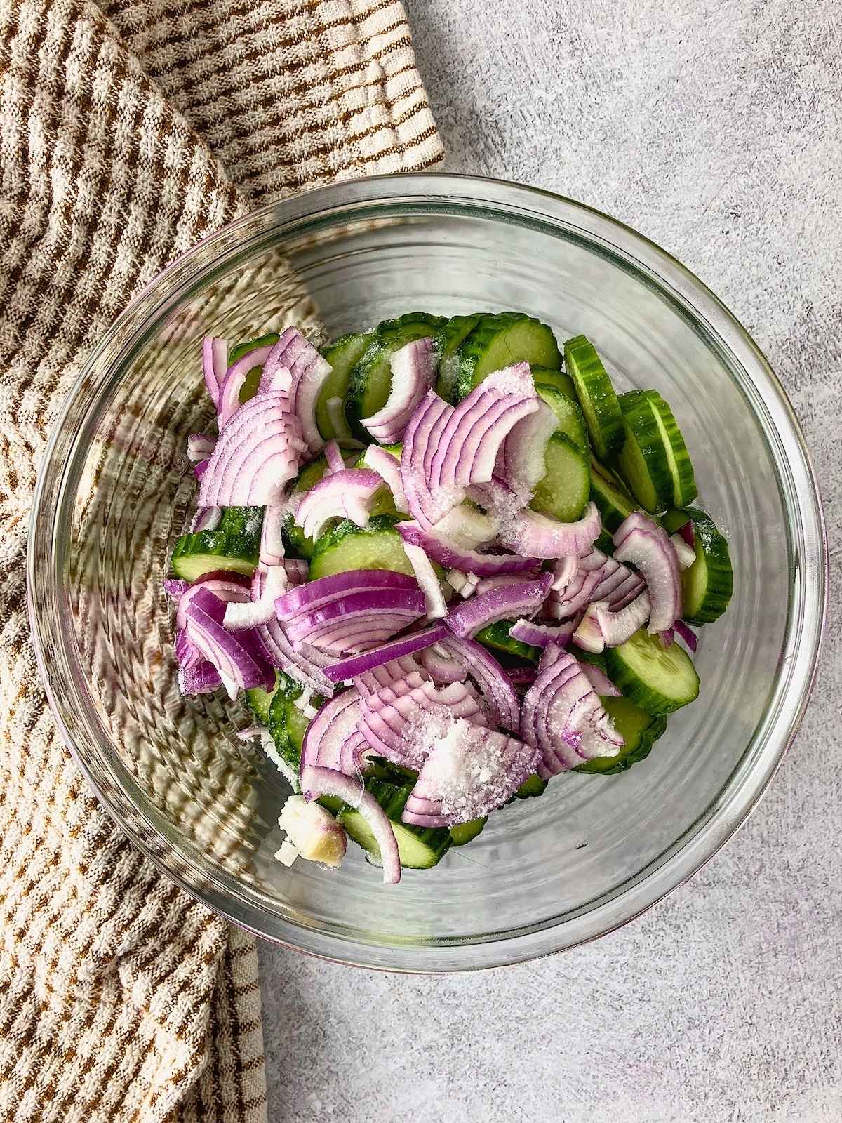 Glass bowl with sliced cucumbers, red onion, and salt ready to be mixed.