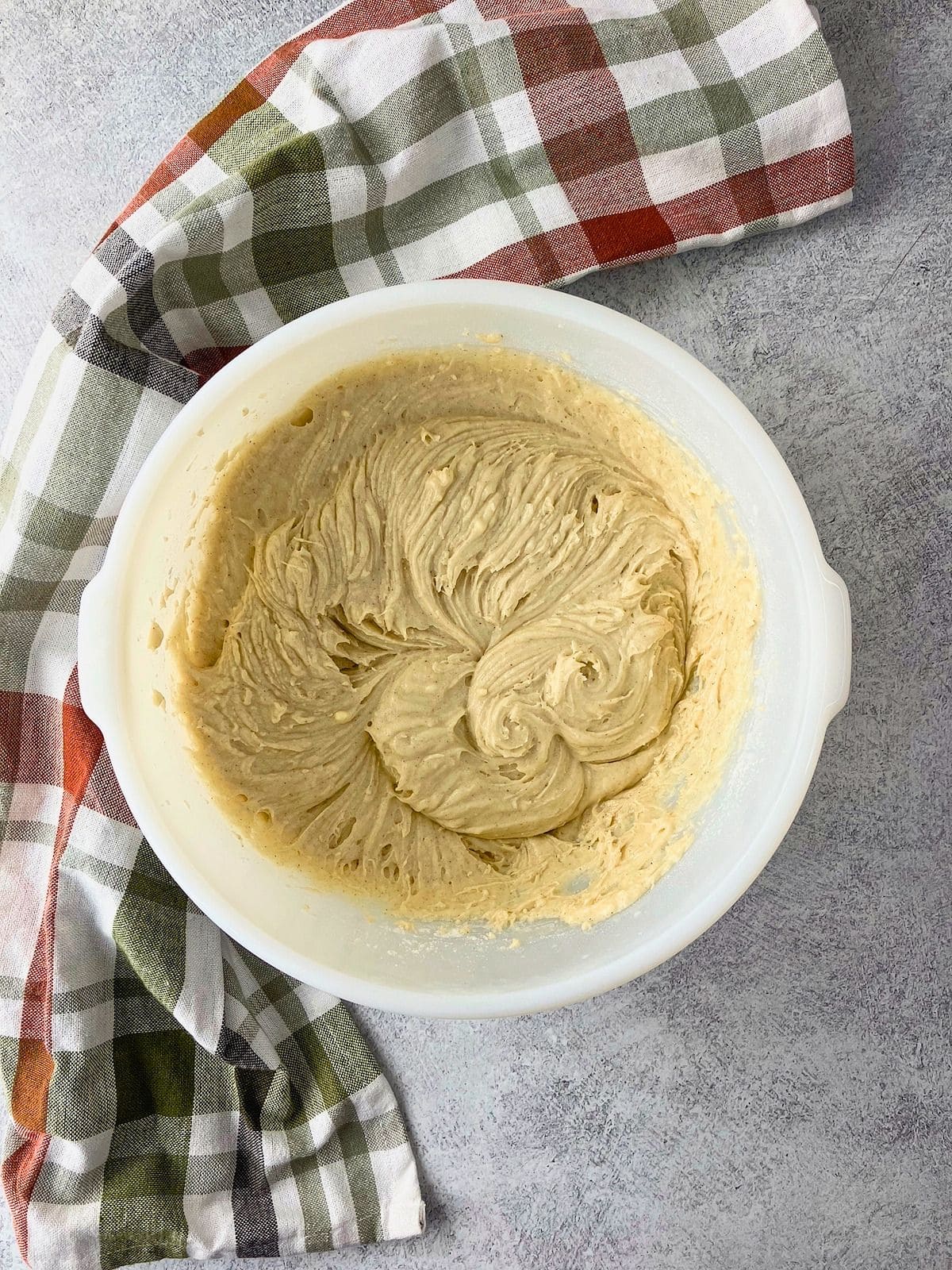 Overhead view of thick pale cobbler batter mixed until smooth and fluffy in a white bowl beside a plaid kitchen towel. The cake topping is ready to be spread over the apples.
