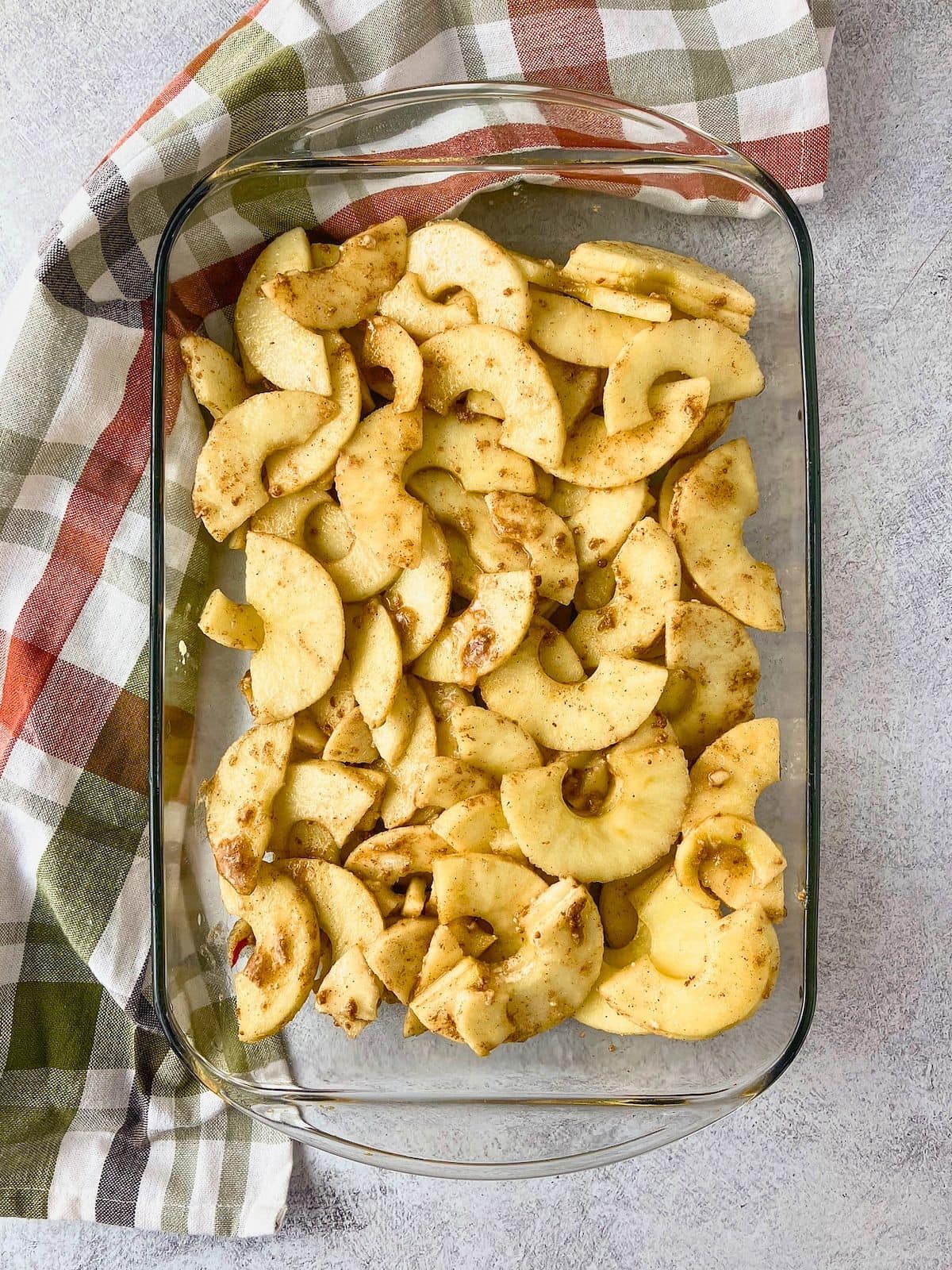 Overhead view of peeled apple slices coated with brown sugar and warm spices spread evenly in a clear glass baking dish.