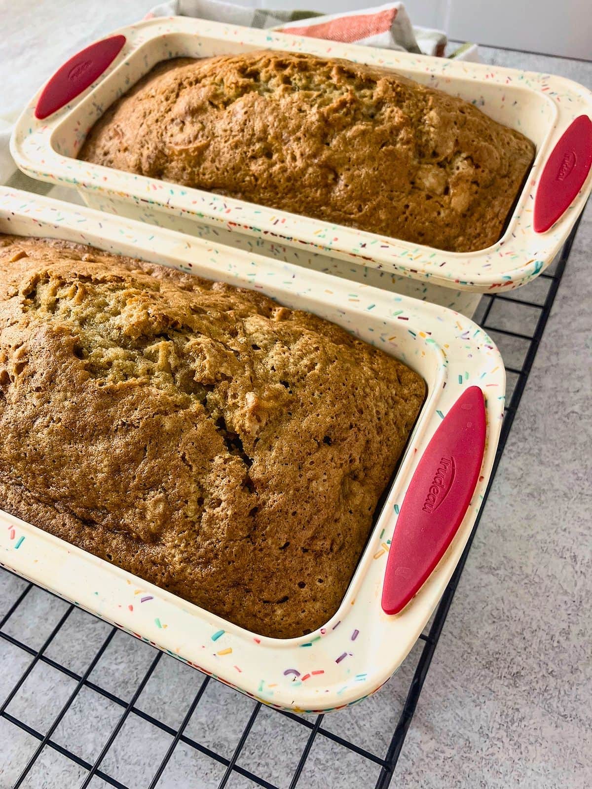 Two freshly baked loaves of apple zucchini bread resting in loaf pans on a wire cooling rack with golden brown cracked tops. The bread is cooling in the pans before being removed and sliced.