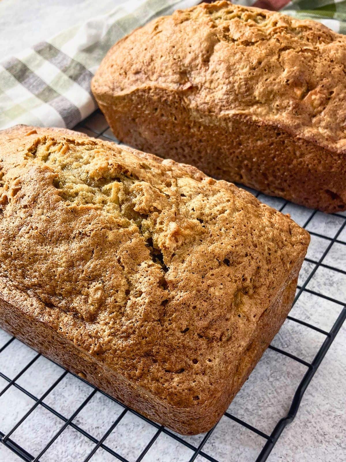 Two freshly baked loaves of apple zucchini bread cooling on a black wire rack with golden brown cracked tops. The loaves are shown cooling before slicing and serving.