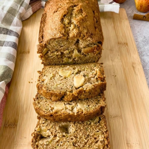 Sliced loaf of apple zucchini bread arranged on a wooden cutting board with visible apple chunks and green zucchini flecks throughout the moist crumb. Whole apples and a cinnamon stick sit in the background beside a plaid kitchen towel.