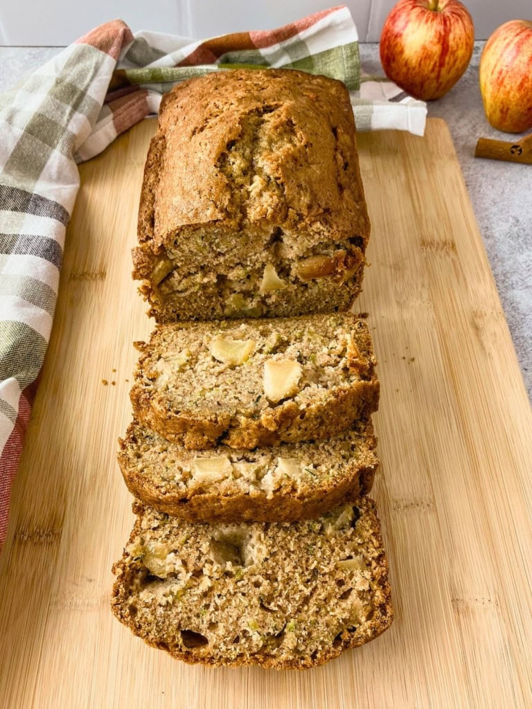 Sliced loaf of apple zucchini bread arranged on a wooden cutting board with visible apple chunks and green zucchini flecks throughout the moist crumb. Whole apples and a cinnamon stick sit in the background beside a plaid kitchen towel.